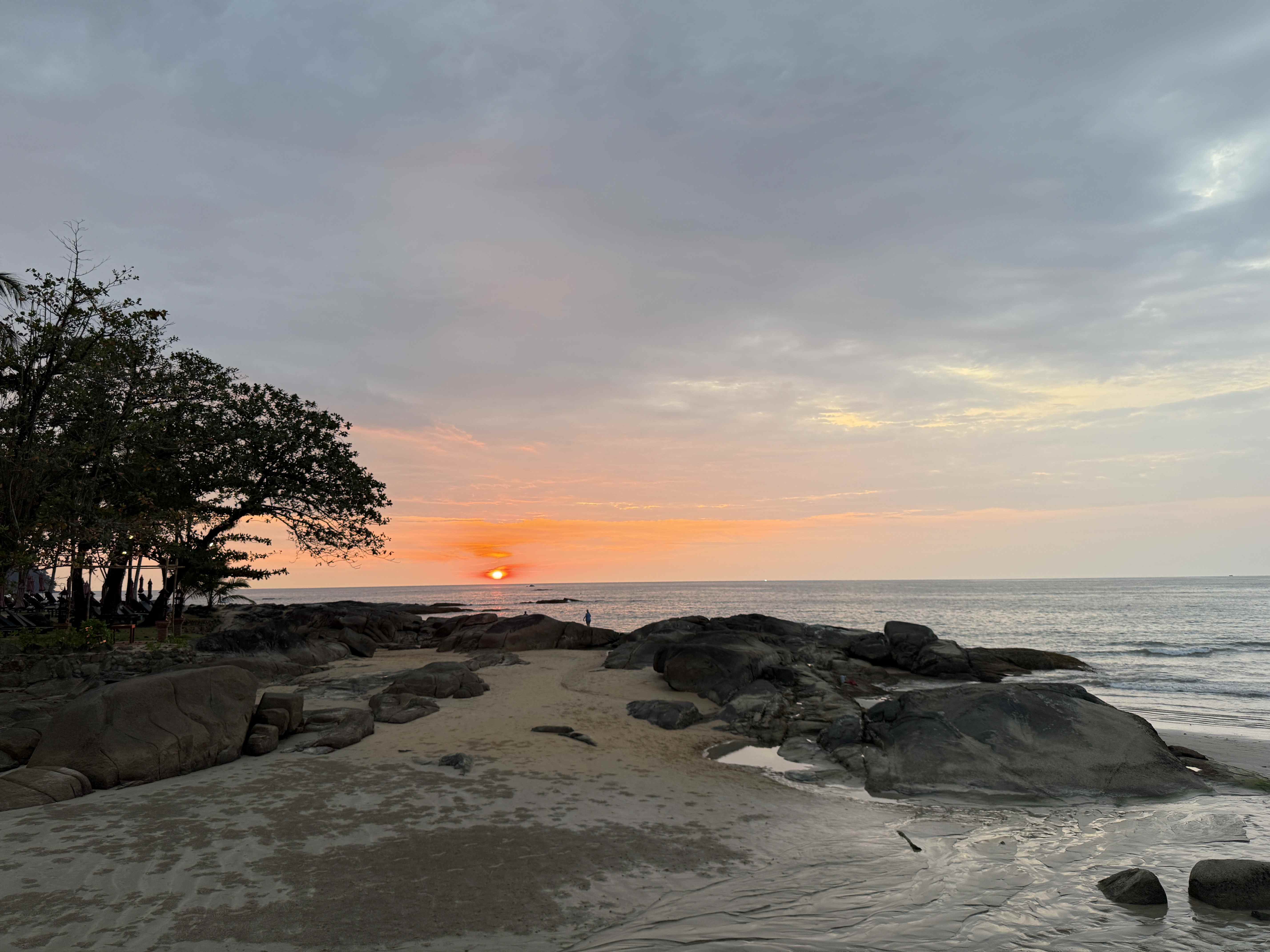 Západ slunce nad mořem, skalnatá pláž s pískem a siluetou stromů