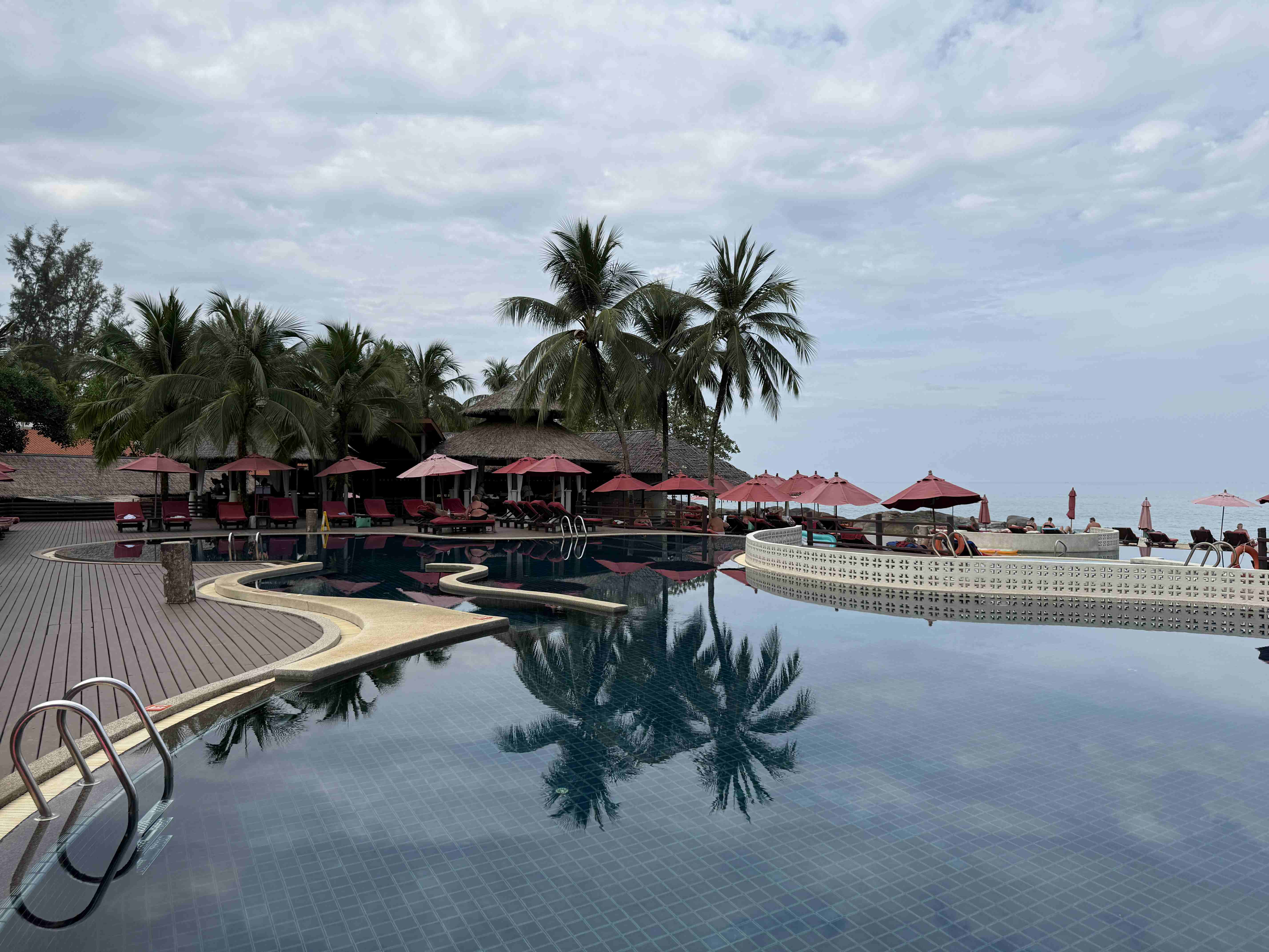 Bazén u pláže s palmami, červenými slunečníky a výhledem na moře