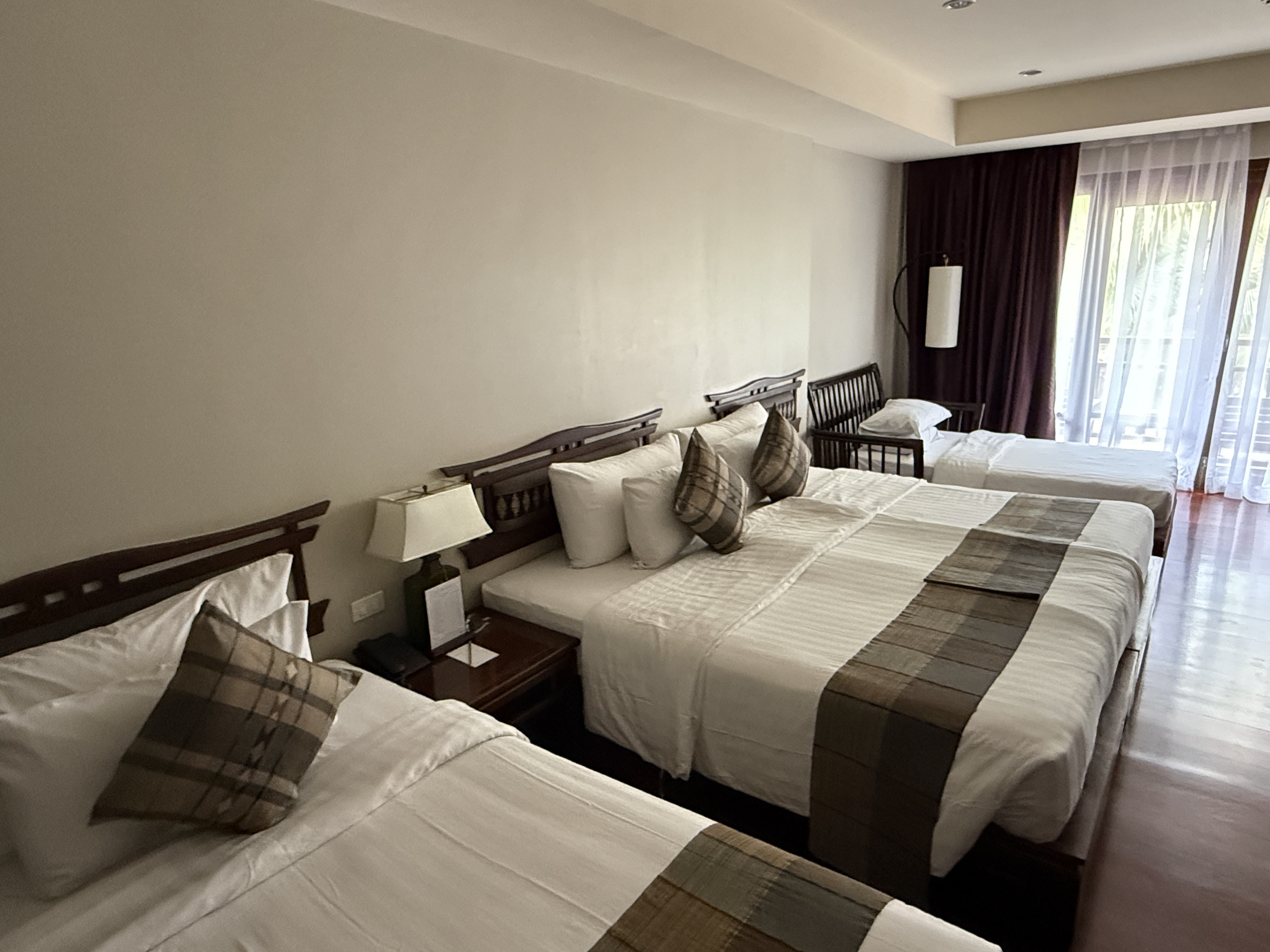 Hotelový pokoj se třemi postelemi, nočním stolkem a balkonem za závěsy