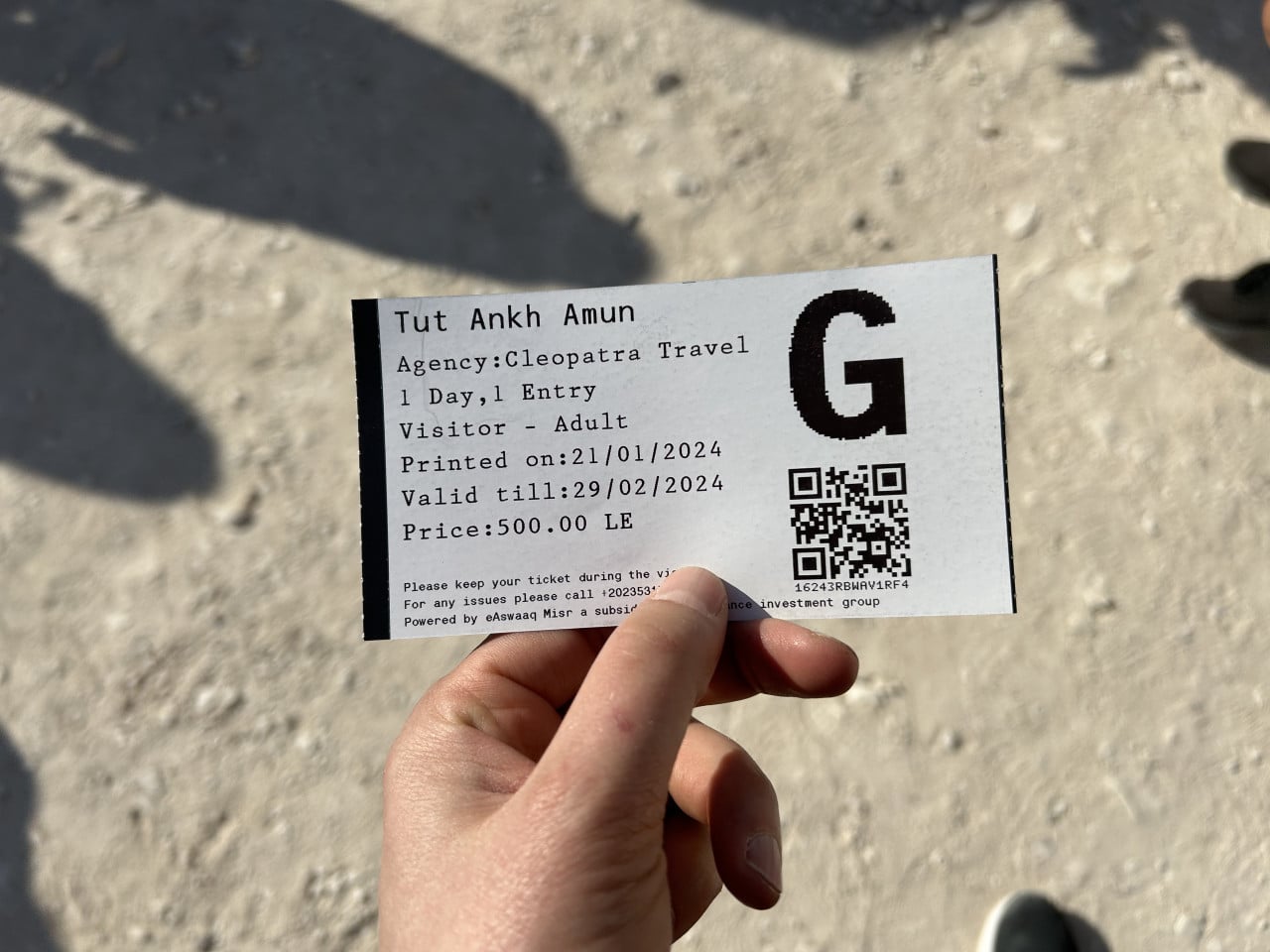 Ruka drží vstupenku Tut Ankh Amun s QR kódem na prašném terénu.