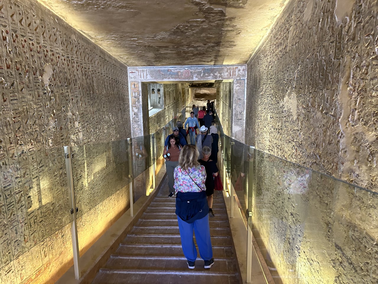Turisté sestupují po schodech v úzké egyptské hrobce s hieroglyfy.