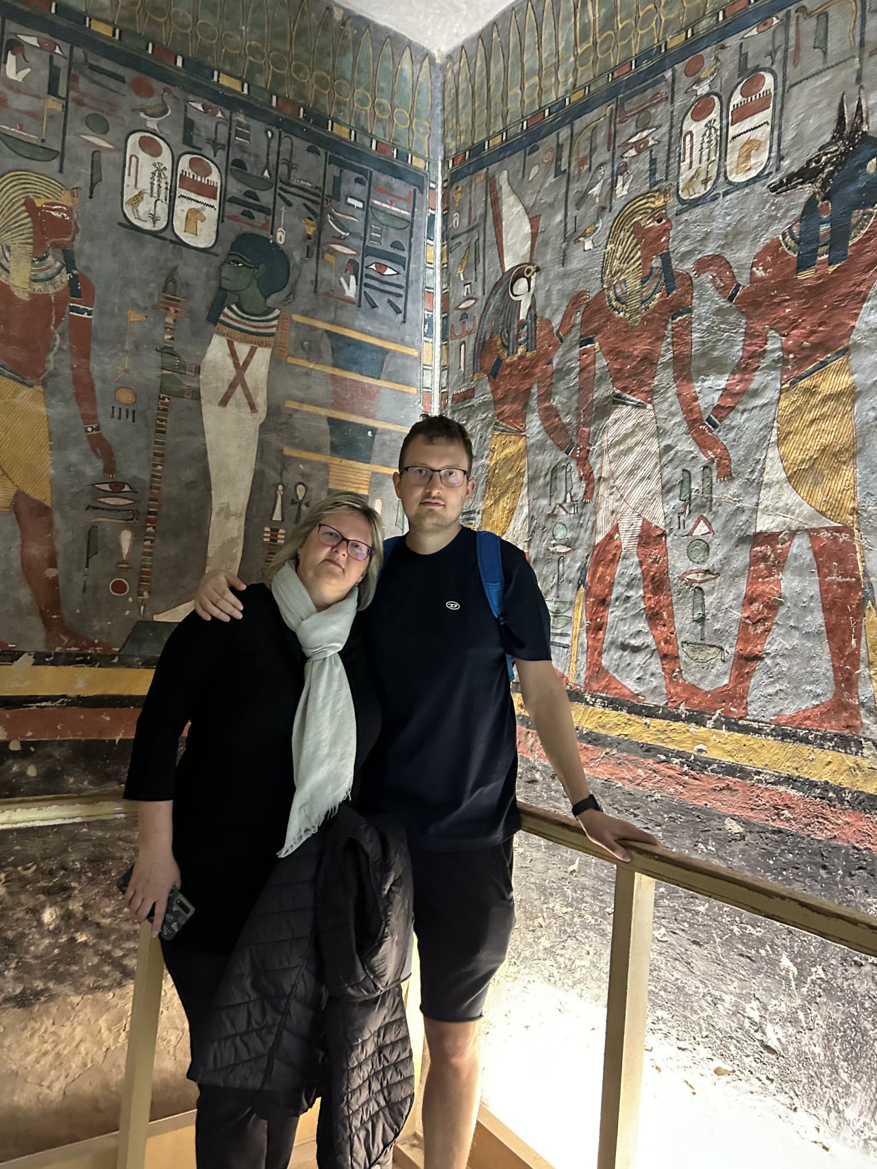 Dva turisté stojí před barevně malovanými egyptskými hieroglyfy v hrobce.