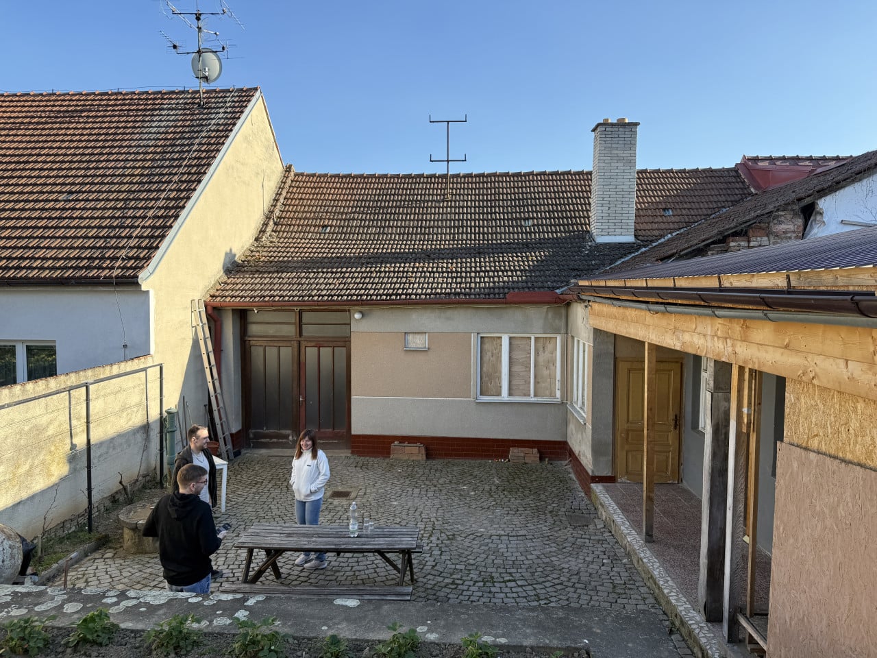 Dvůr s několika lidmi, starými střechami, žebříkem a dřevěným přístřeškem.