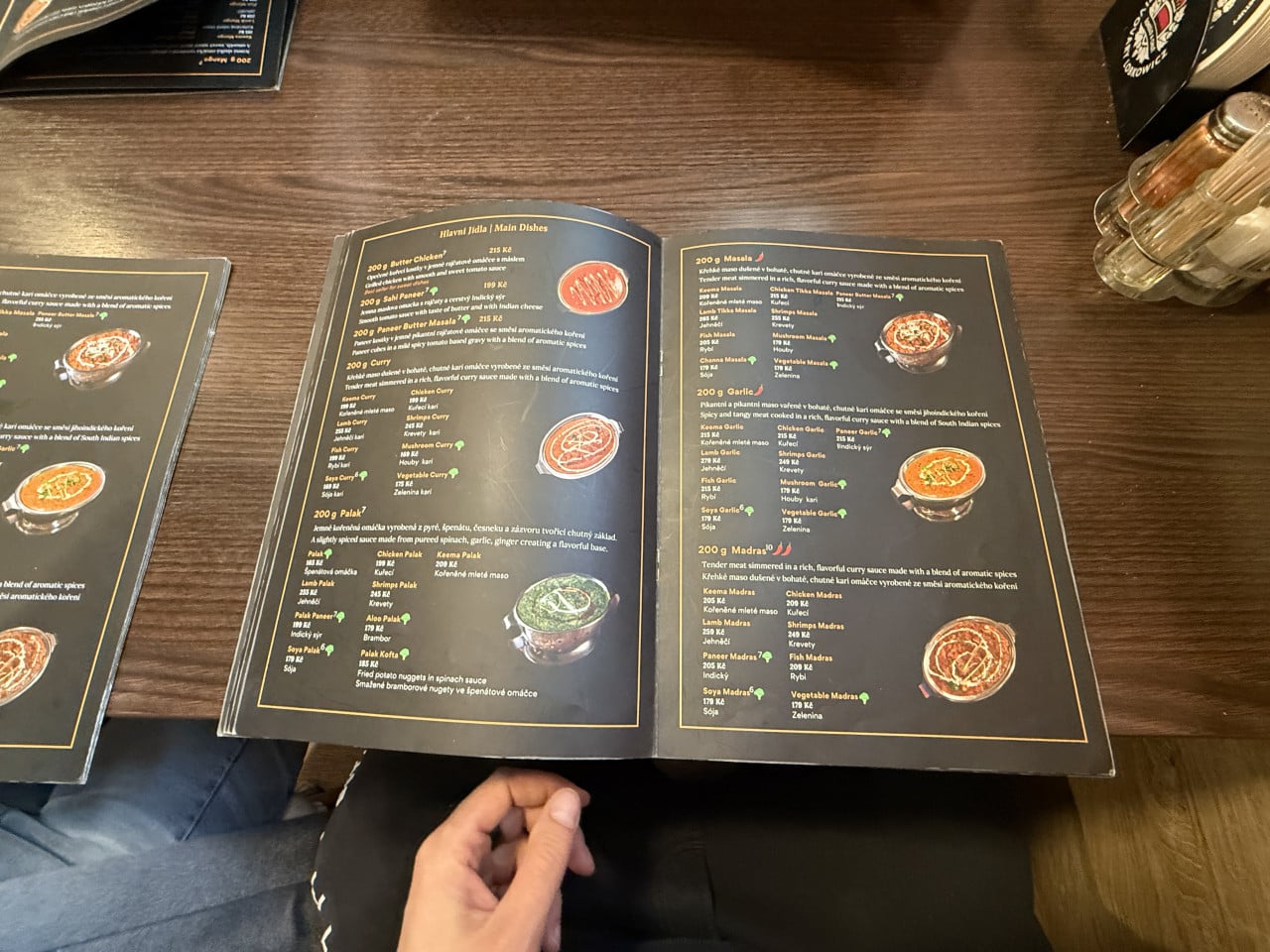 Otevřené menu s indickými jídly na dřevěném stole, ruce drží stránku.