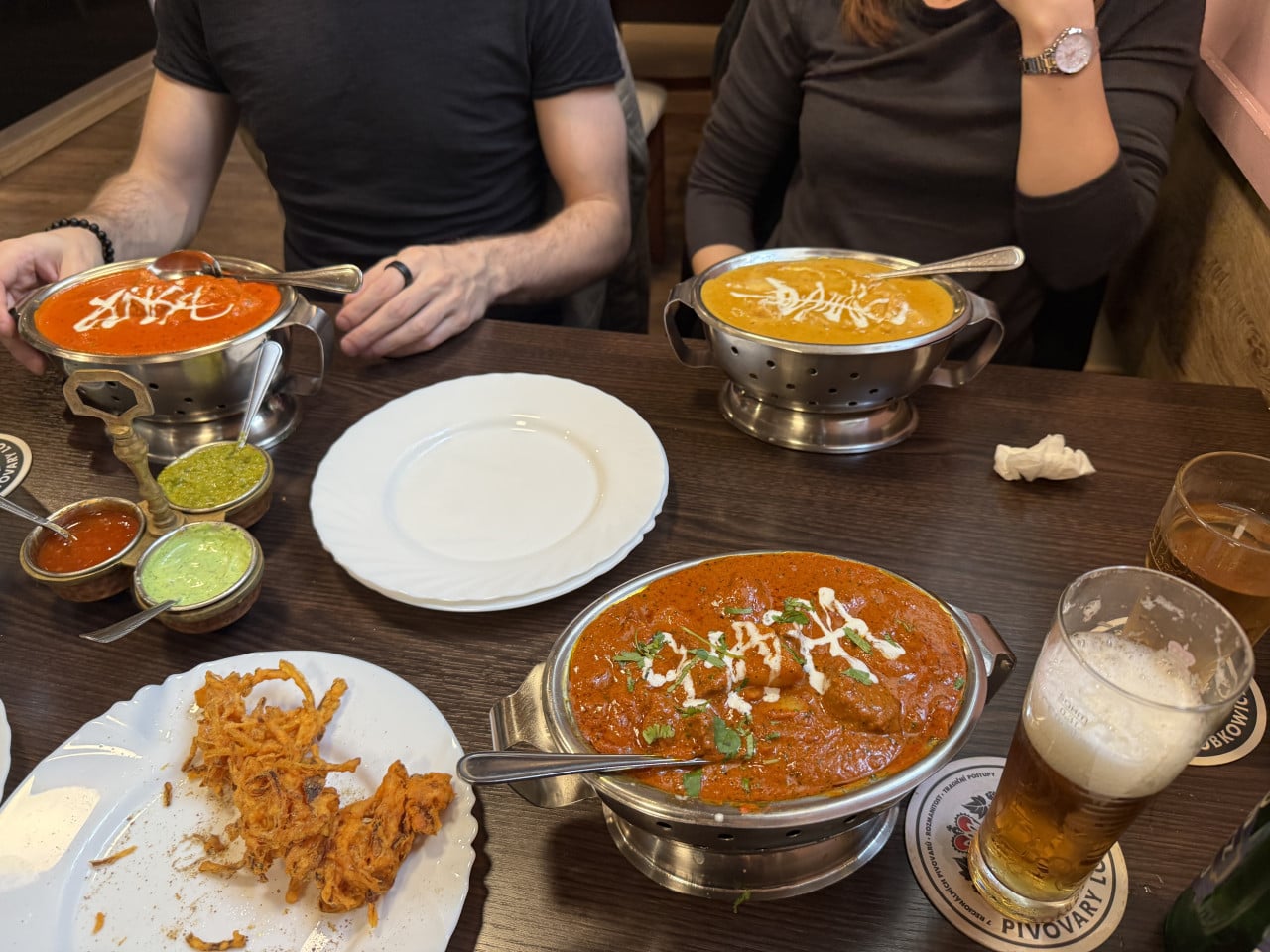 Dva lidé sedí u stolu s indickým jídlem, omáčkami a pivy.
