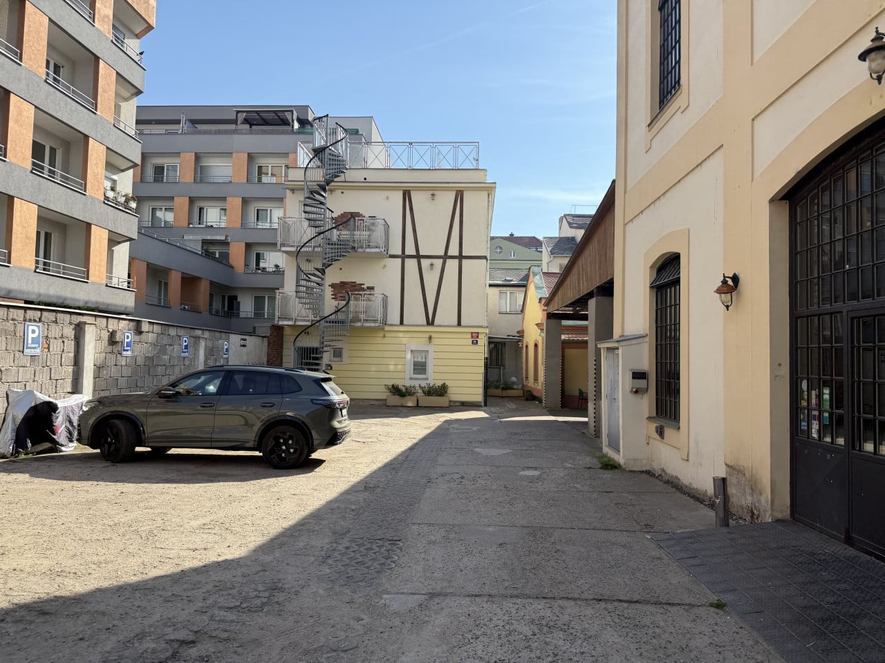 Dvůr mezi budovami s točitým schodištěm, zaparkovaným SUV a modrou oblohou.