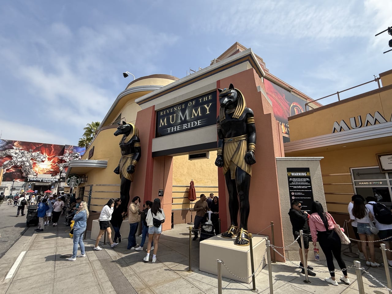 Vstup k atrakci Revenge of the Mummy The Ride s černými sochami Anubise a návštěvníky v řadě