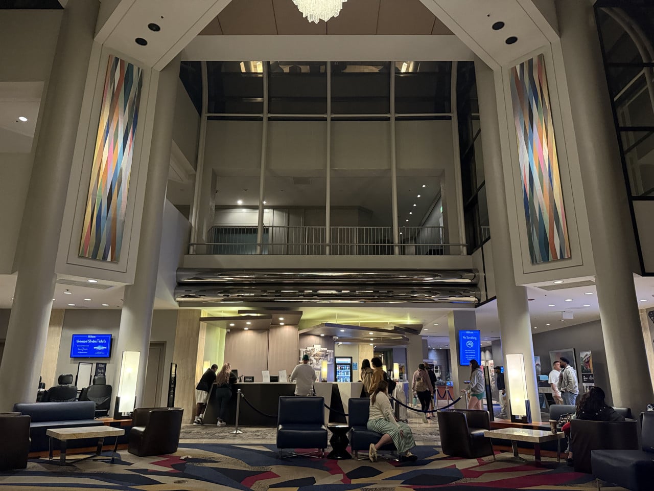 Hotelová lobby s recepcí, barevnými nástěnnými panely a hosty čekajícími u přepážky