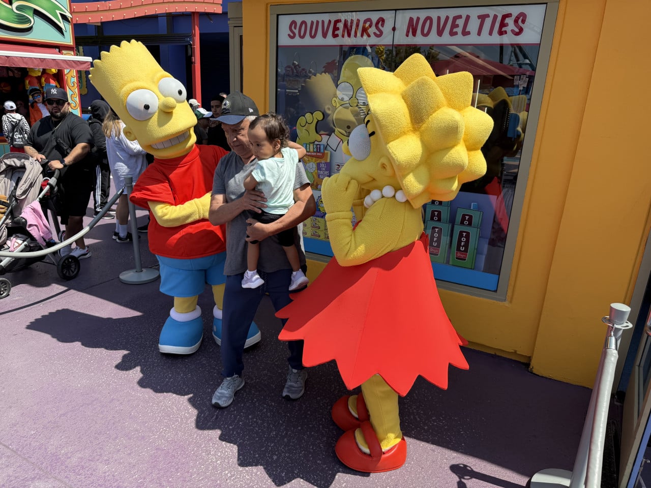 Bart Simpson a Lisa Simpson pózují s mužem a dítětem u obchodu se suvenýry.