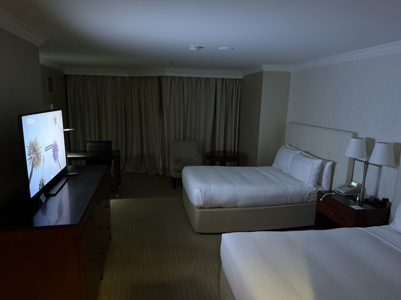 Hotelový pokoj Hilton s dvěma postelemi, televizí a zataženými závěsy.