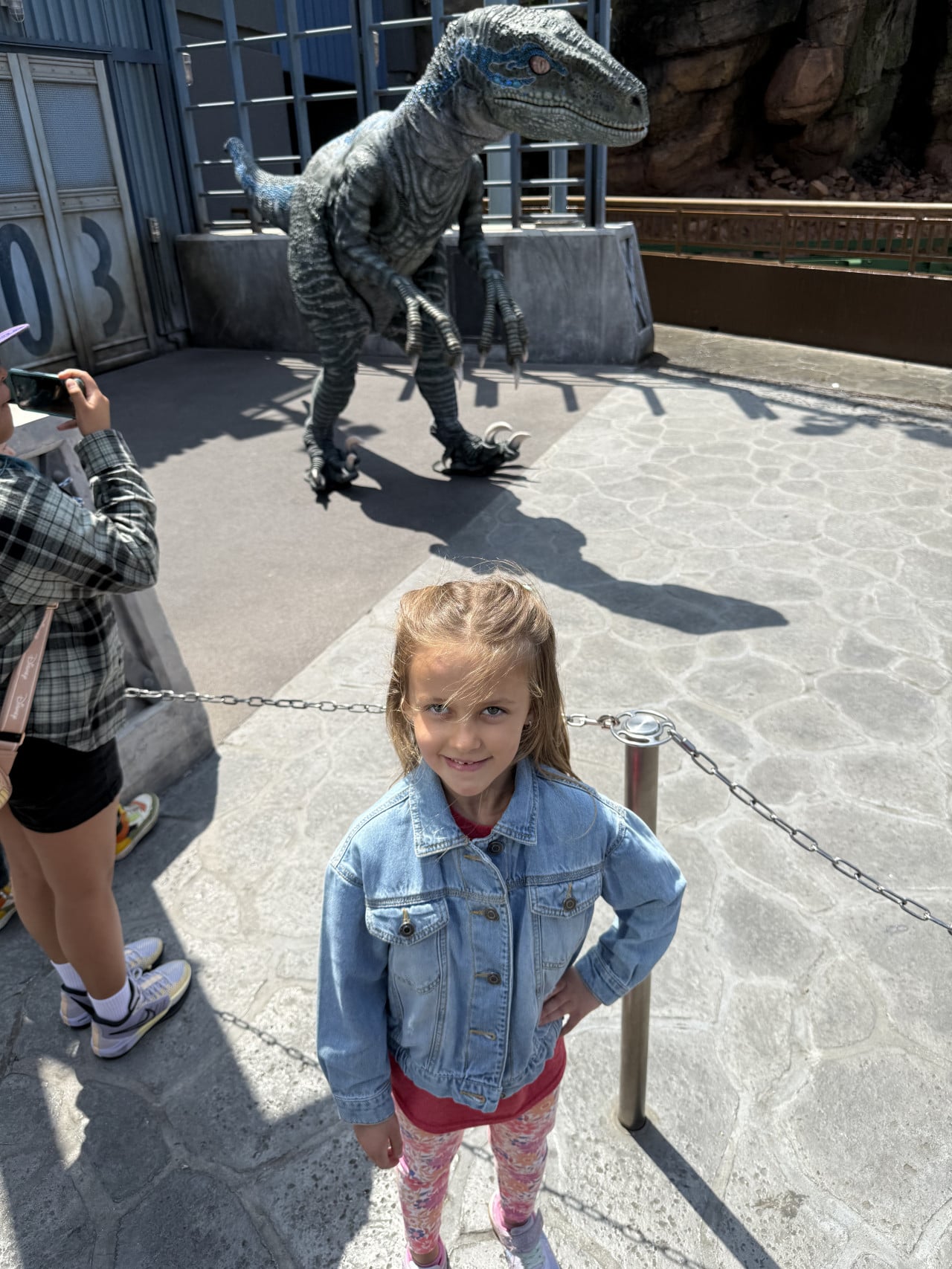 Dívka v džínové bundě stojí před dinosaurím animatronikem v Universal Studios Hollywood.
