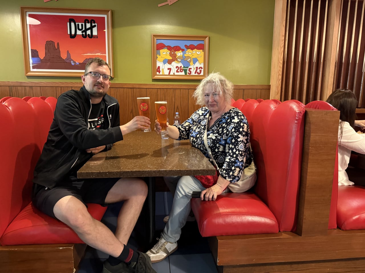 Dva lidé v restauraci se Simpsonovými, připíjejí si půllitry Duff Beer.