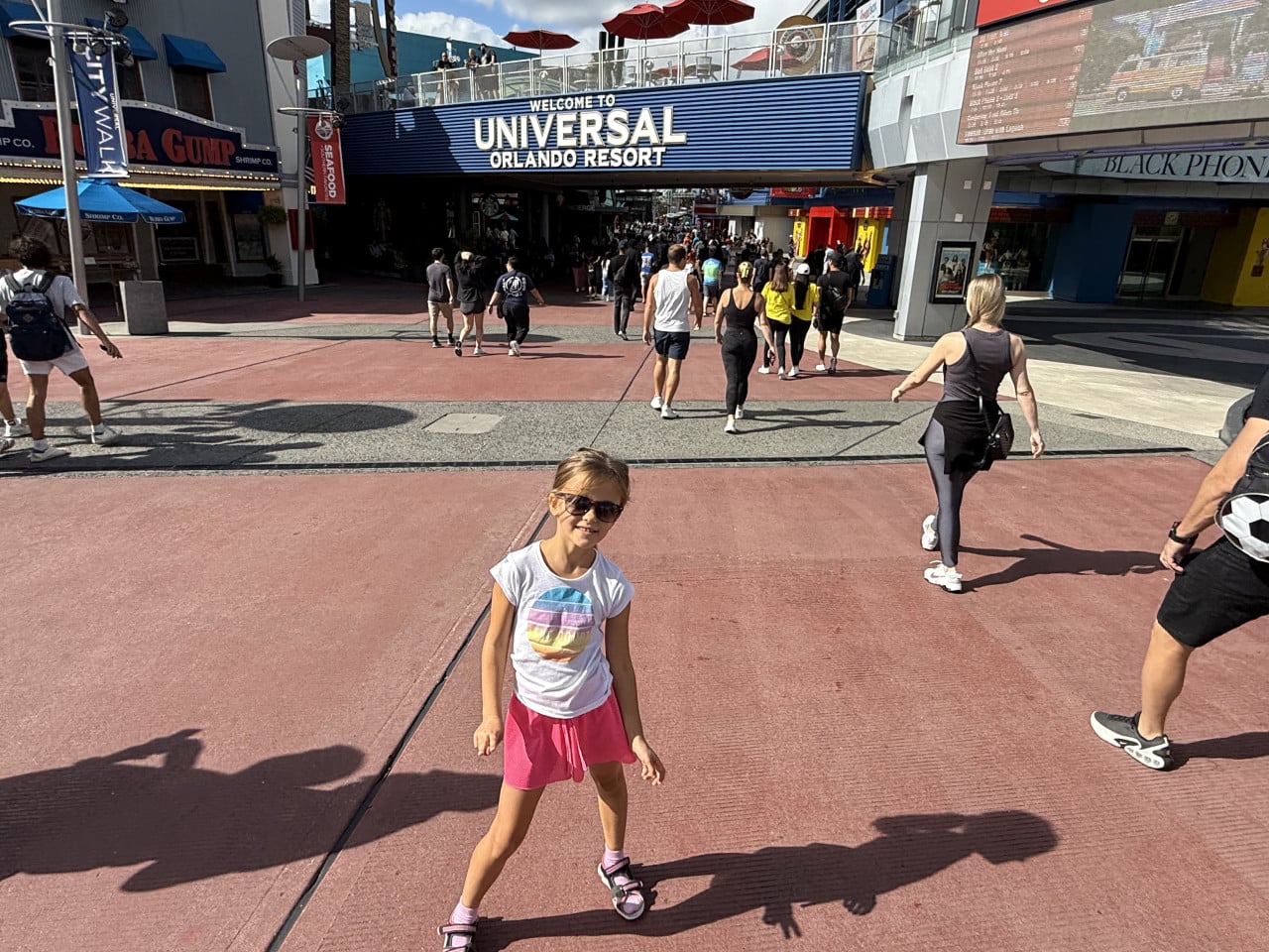 Dívka v brýlích stojí před nápisem Welcome to Universal Orlando Resort, lidé kolem procházejí parkem.