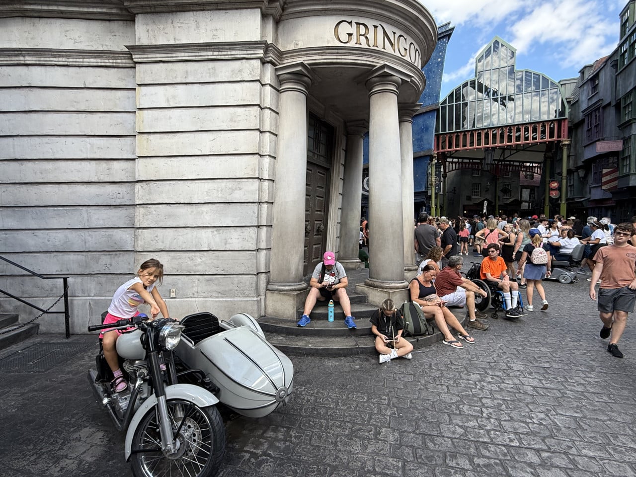 Dívka na motorce s postranním vozíkem před budovou Gringottovy banky, kolem čekají návštěvníci Universal Studios Orlando