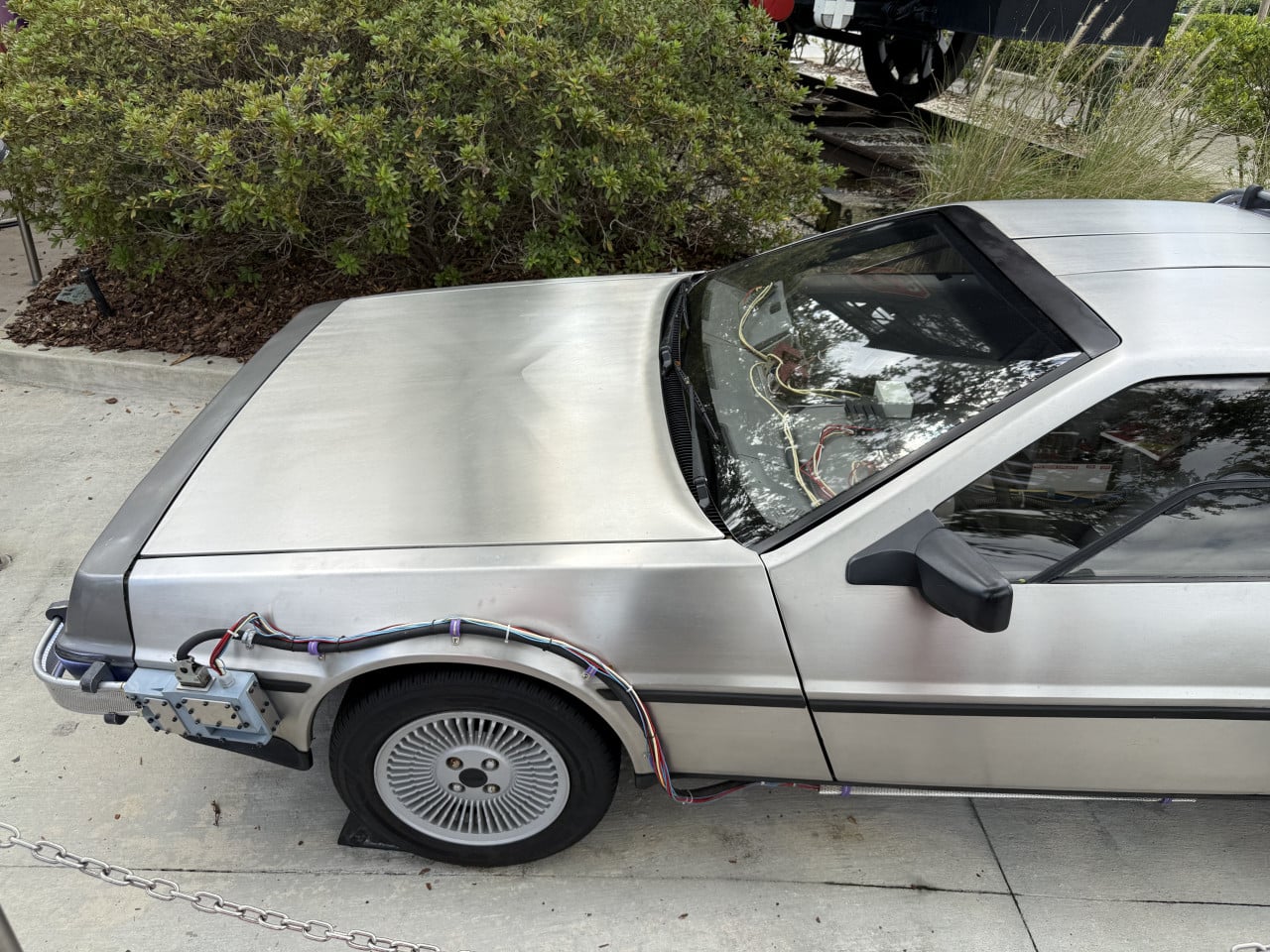 Stříbrný DeLorean s odkrytými kabely, zaparkovaný u chodníku mezi keři.