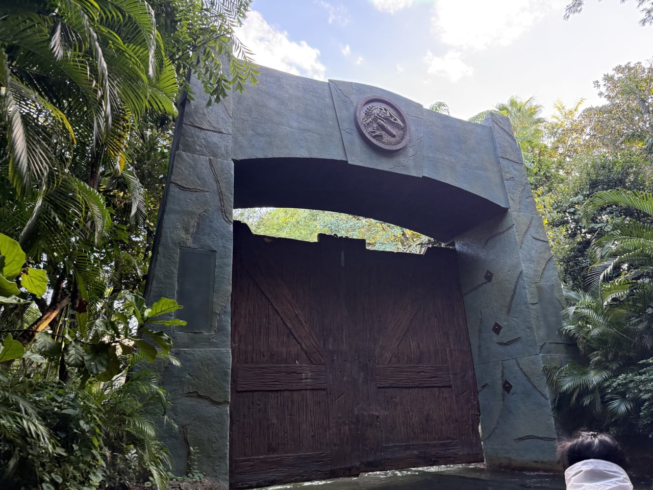 Vstup do dinoparku s kamennou branou, dřevěnými vraty a tropickou zelení.