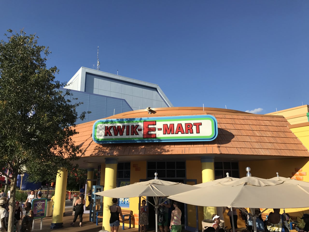 Kwik-E-Mart v Universal Studios s návštěvníky a slunečníky pod modrou oblohou