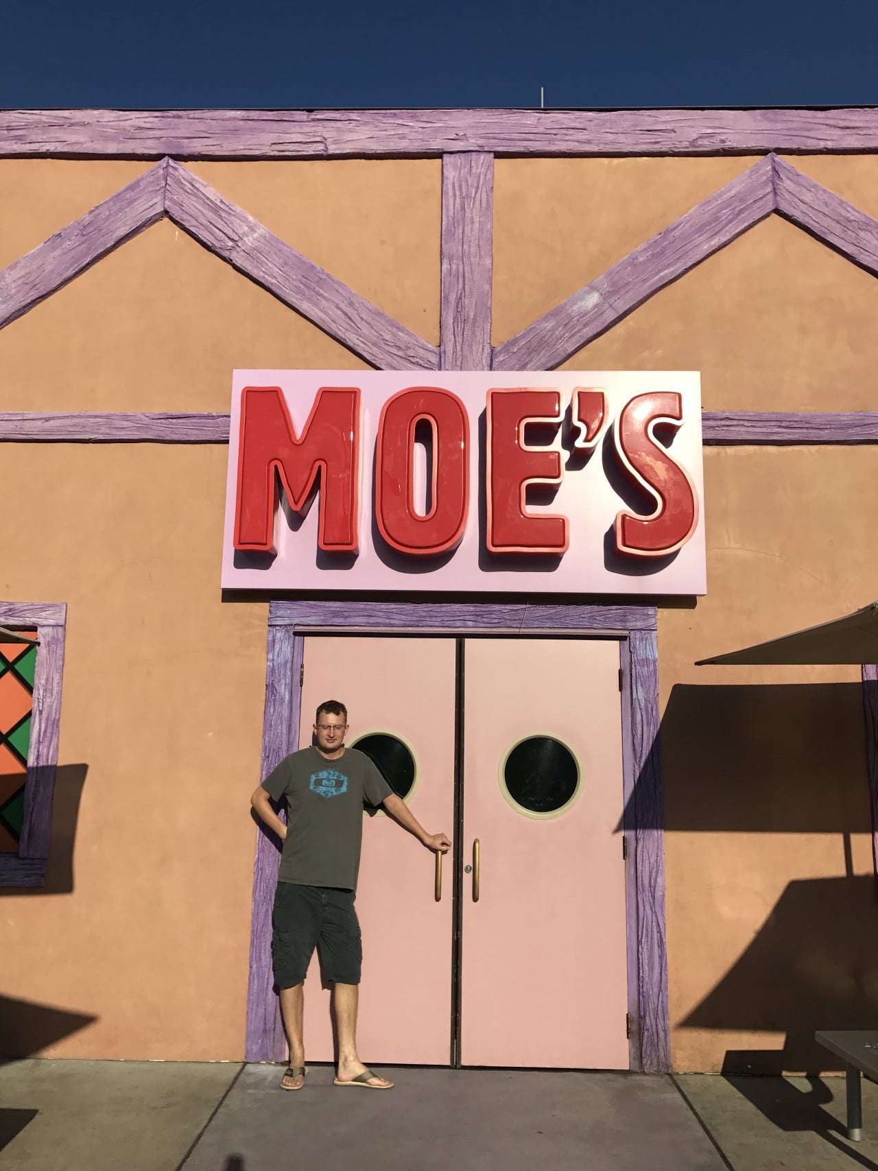 Muž stojí před pastelovou fasádou restaurace Moe's s velkým červeným nápisem.
