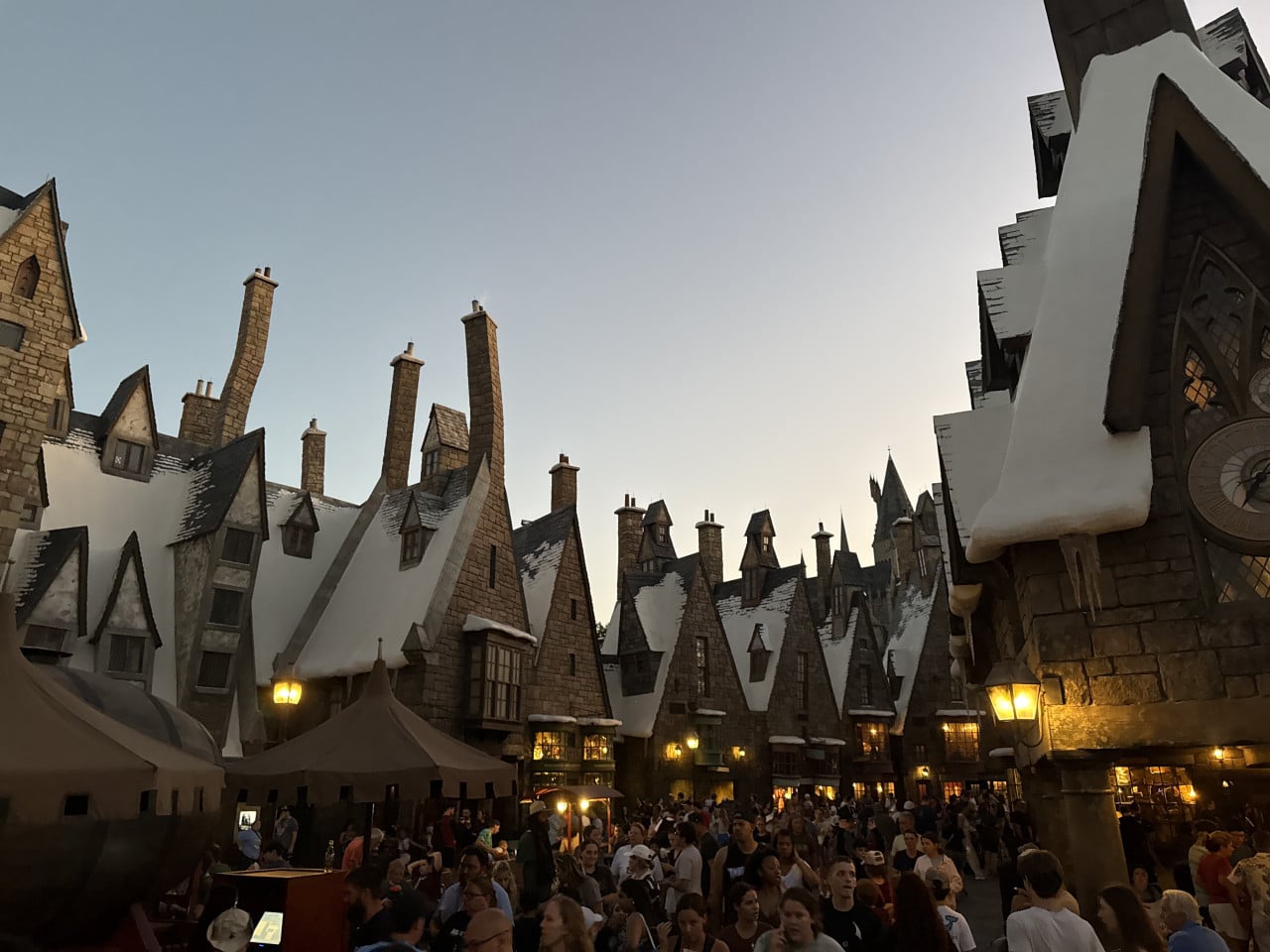 Večerní kouzelnická vesnice s zasněženými střechami, komíny a davem návštěvníků v Universal Studios Orlando