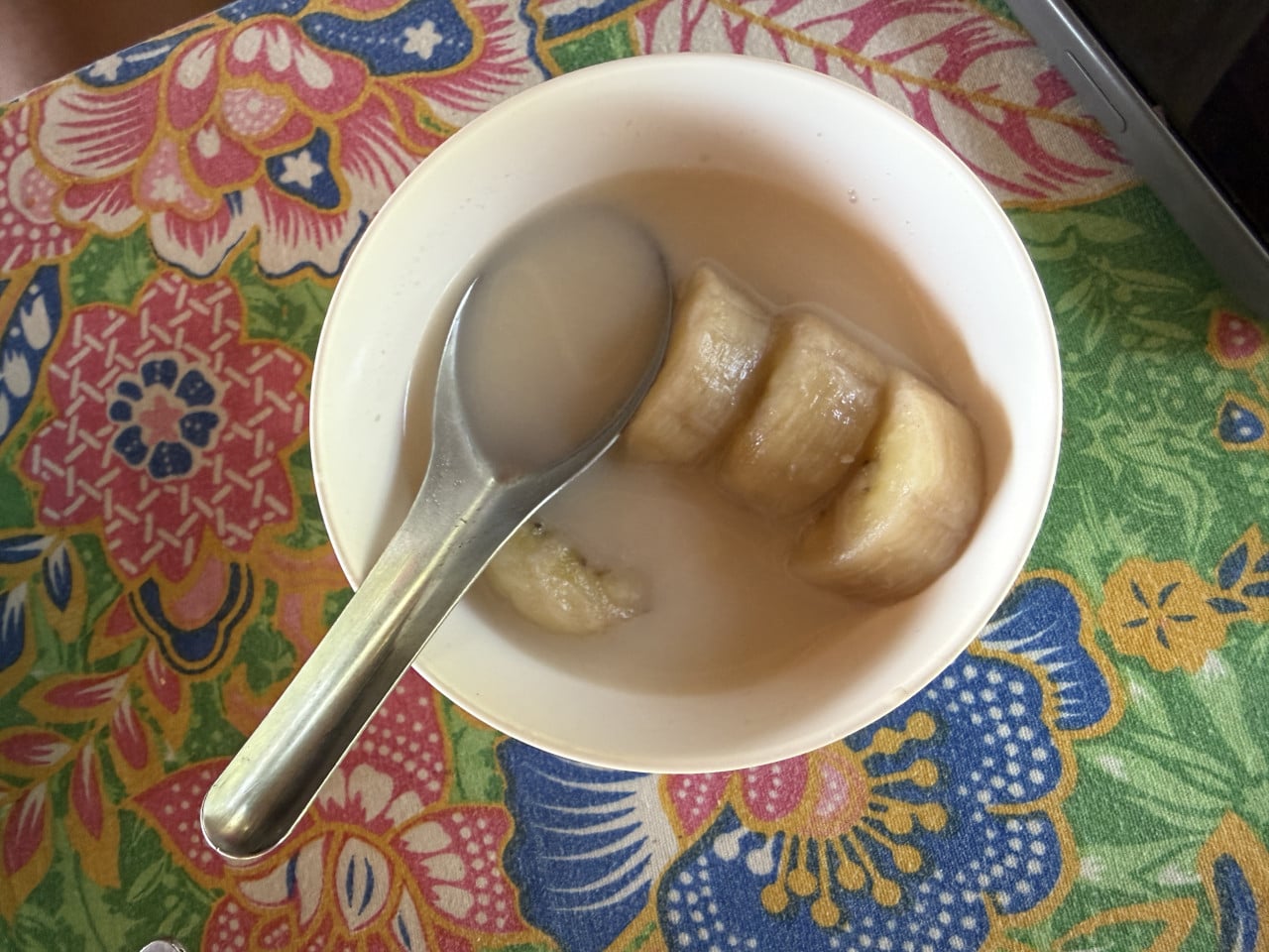 Bílá miska s banánem v kokosovém mléce a lžící na květovaném ubrusu.