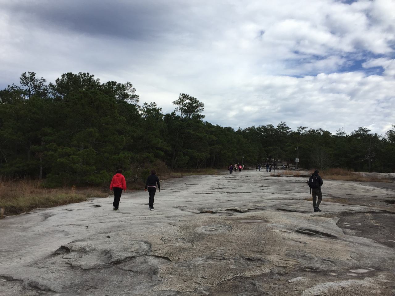 Turisté kráčejí po žulové plošině Stone Mountain, kolem les, zataženo