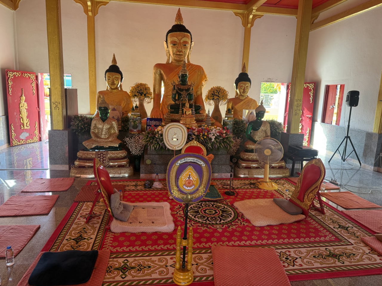 Interiér chrámu s velkými sochami Buddhy, květinami, thajskými nápisy a modlitebními rohožemi.
