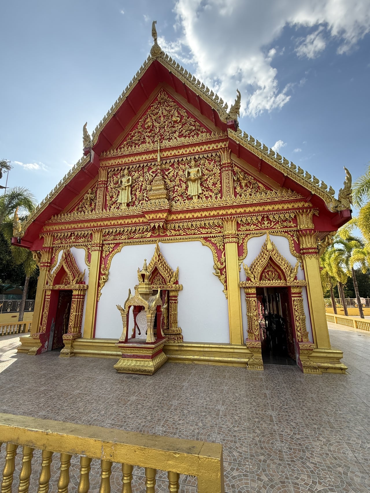 Zdobená červeno-zlatá fasáda thajského chrámu s otevřeným vstupem, palmy, modrá obloha