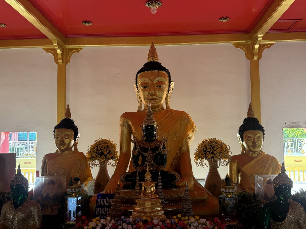 Velká zlatá socha sedícího Buddhy v chrámu, po stranách menší, květiny, červený strop.