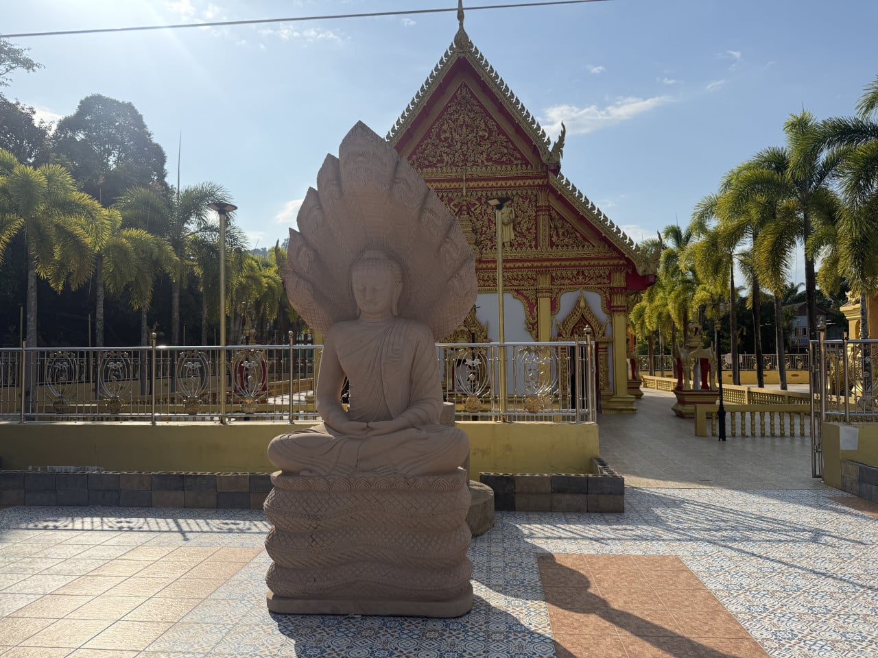 Kamenná socha Buddhy s hadem naga před zdobeným chrámem a palmami