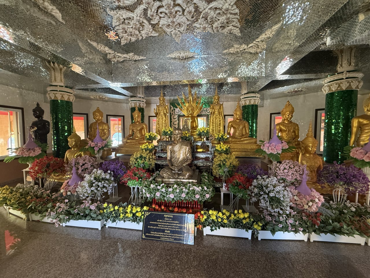 Oltář s pozlacenými sochami Buddhy, sochou mnicha, květinami a thajským nápisem.