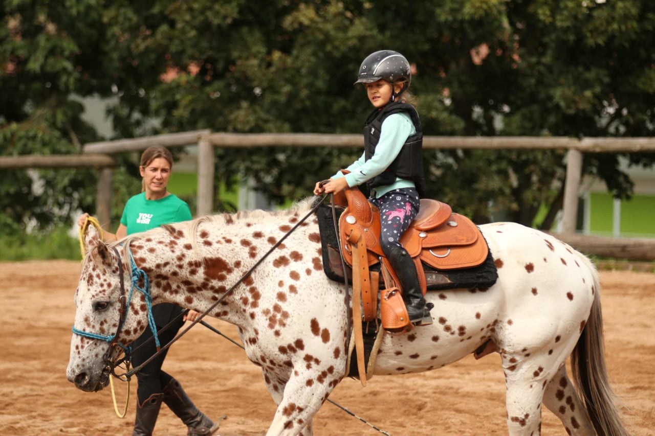 Dívka v helmě jede na strakatém koni ve westernovém sedle, trenérka s tričkem you.