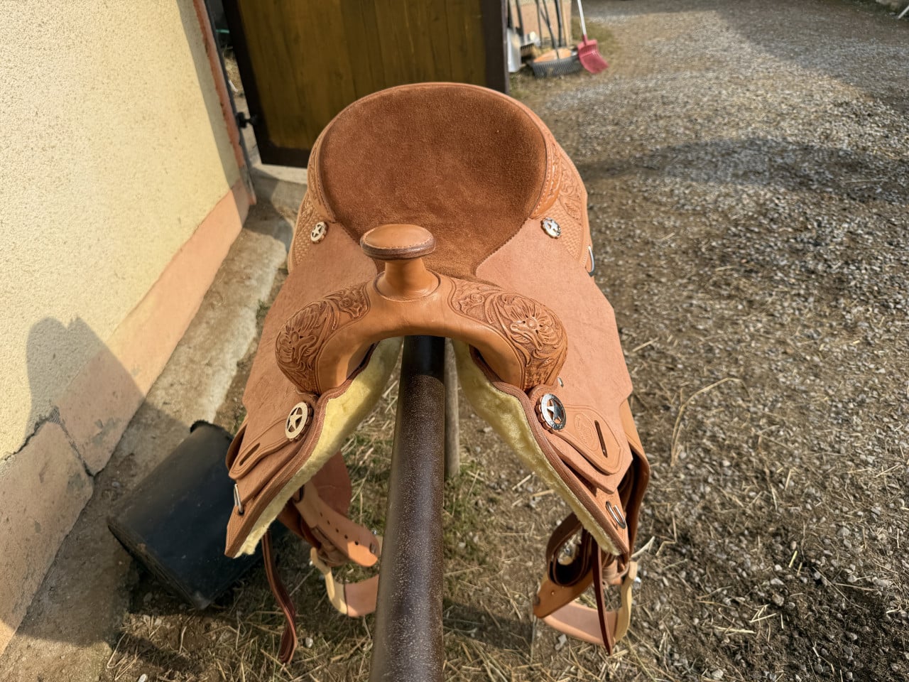 Detail koženého westernového sedla s vyřezávaným zdobením položeného na stojanu u stáje.