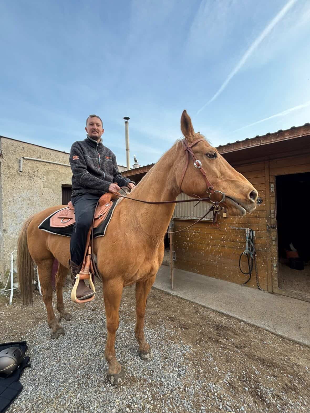 Muž sedí na koni s westernovým sedlem na dvoře u stájí