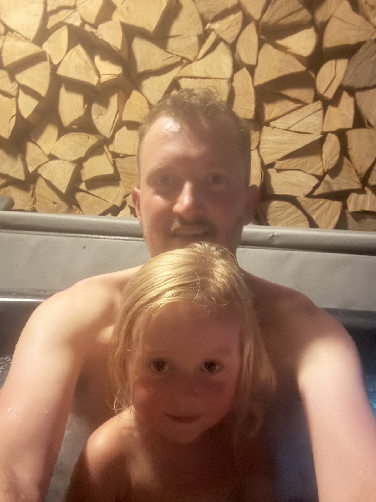 Muž a malá holčička ve vířivce pod stěnou z naskládaného dřeva