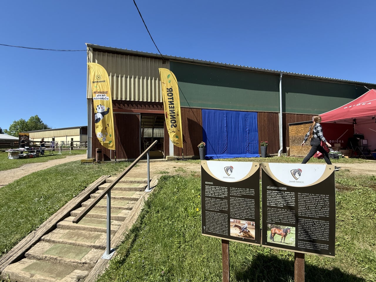 Areál Smart Ranch s informačními tabulemi, žlutými vlajkami a modrým závěsem u haly.