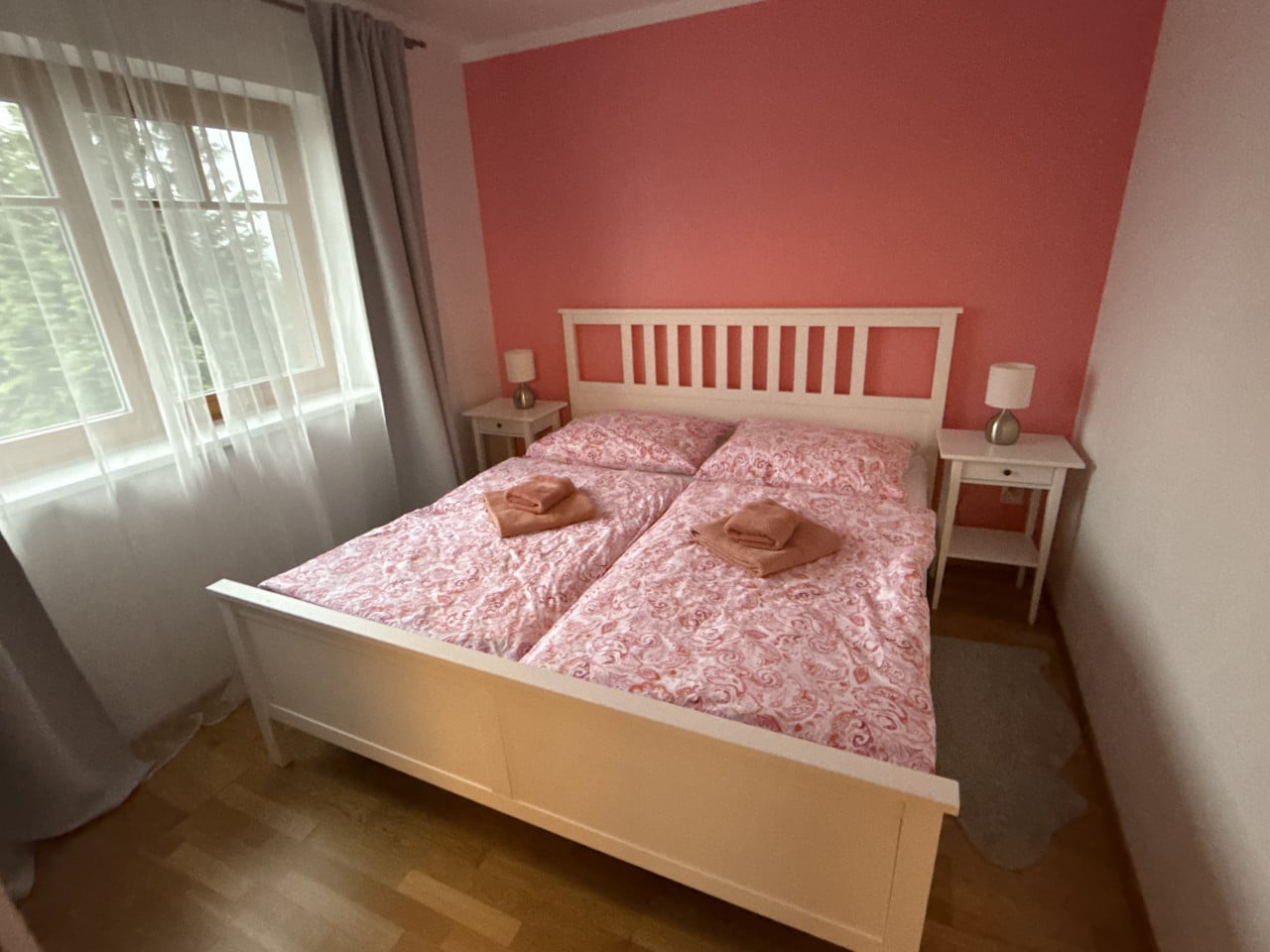 Světle růžová ložnice s manželskou postelí, nočními stolky a oknem se závěsy.