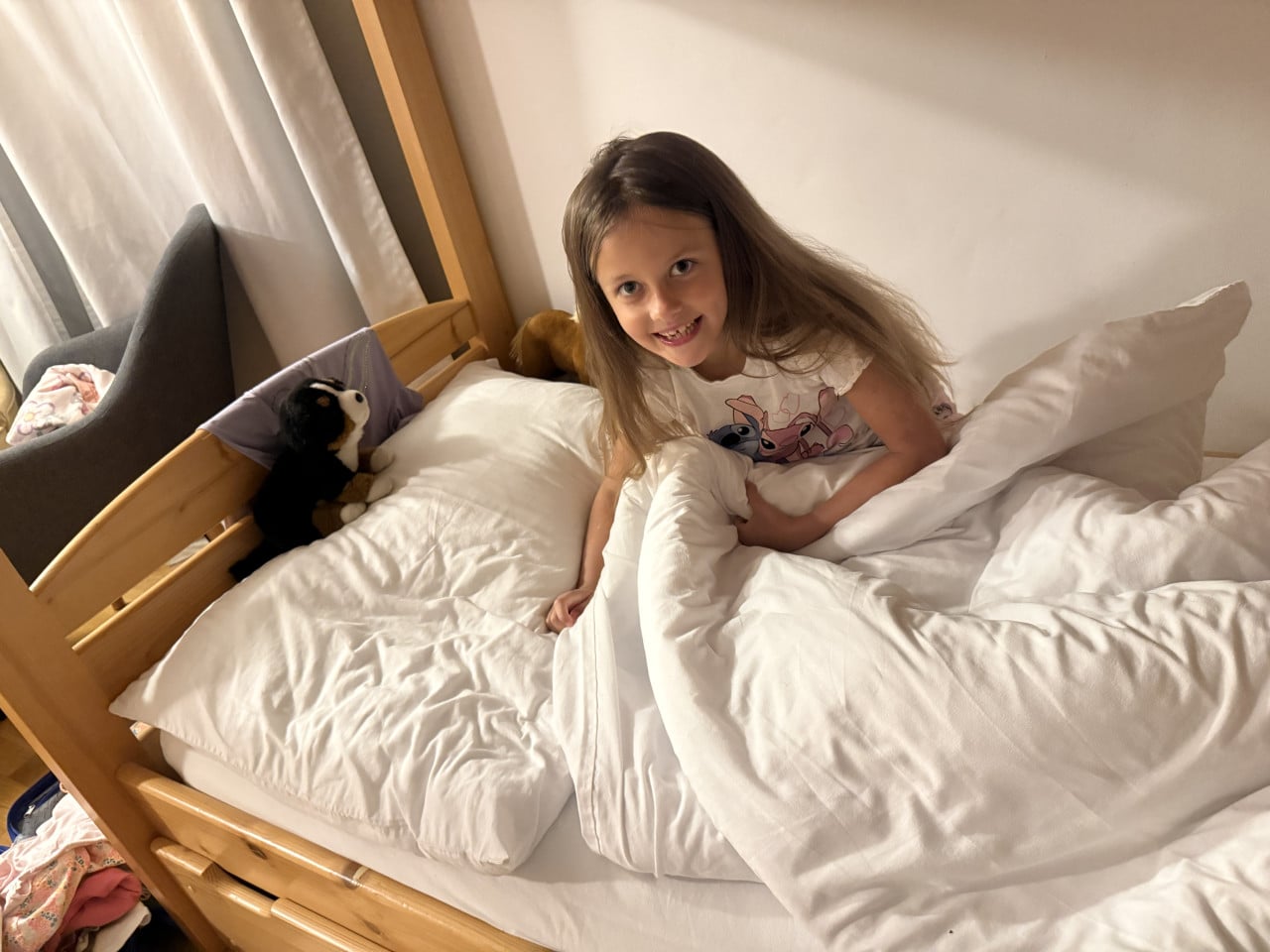 Usměvavá holčička sedí v posteli s bílými peřinami a plyšovým psem.