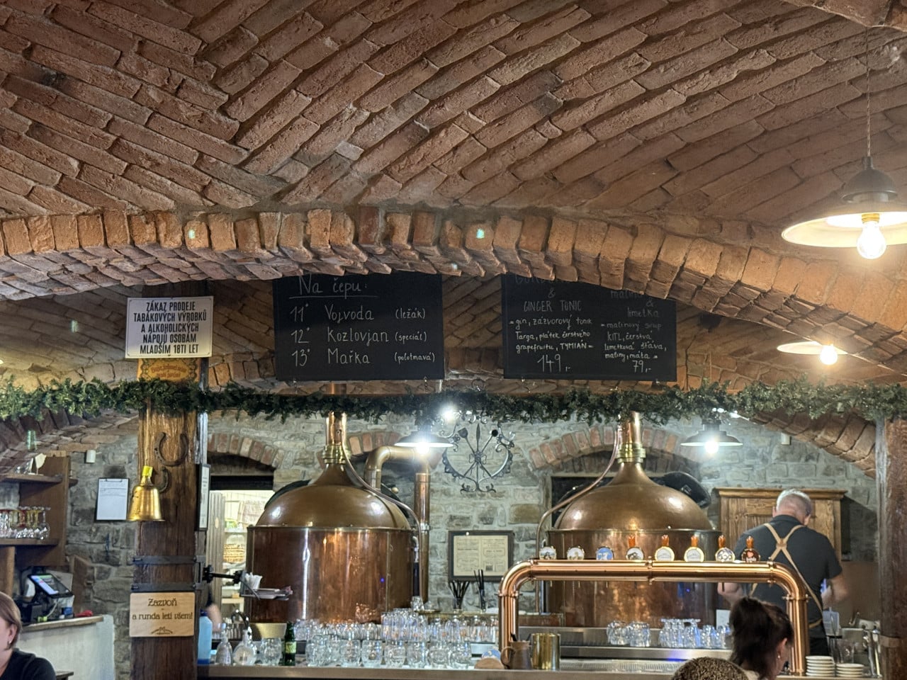 Rustikální bar s měděnými tanky, tabulemi s nápoji a cihlovou klenbou.