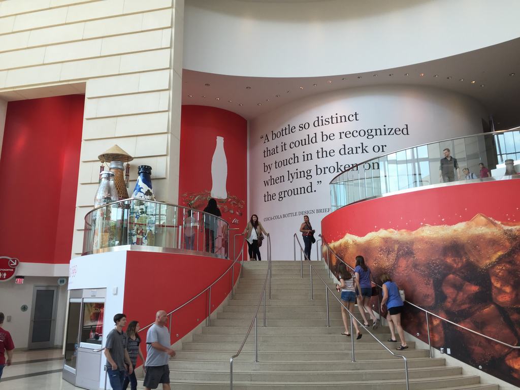 Vstupní hala World of Coca‑Cola, schody, lidé a citát o lahvi na zdi