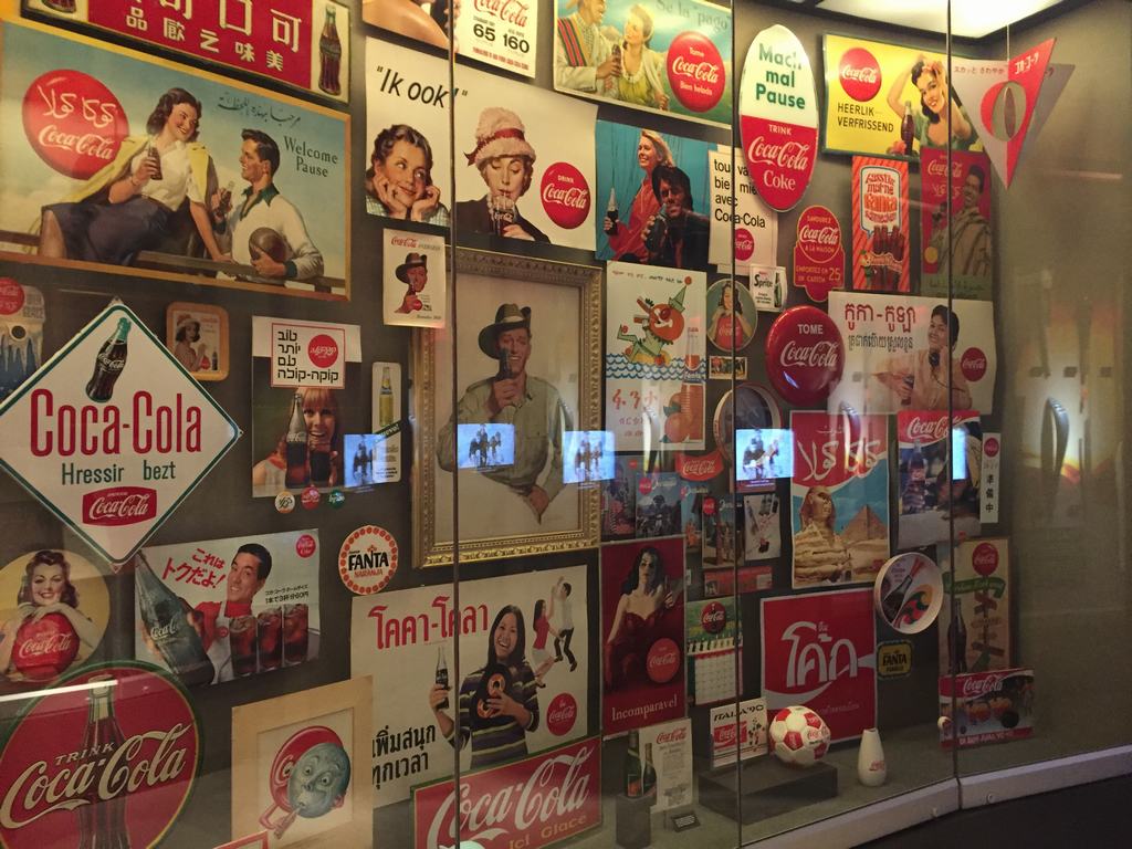 Stěna s vintage plakáty a reklamami Coca‑Cola, nápisy Coca‑Cola a Drink Coca‑Cola