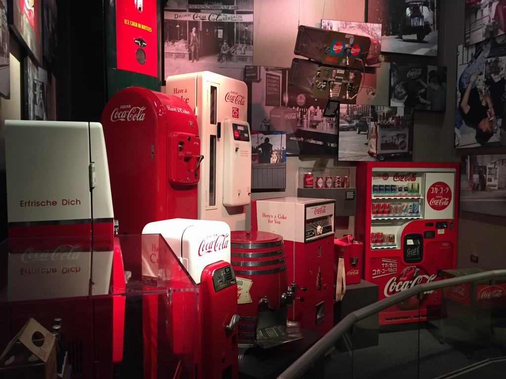 Expozice s retro automaty Coca‑Cola; nápisy Erfrische Dich a Here’s a Coke for You.