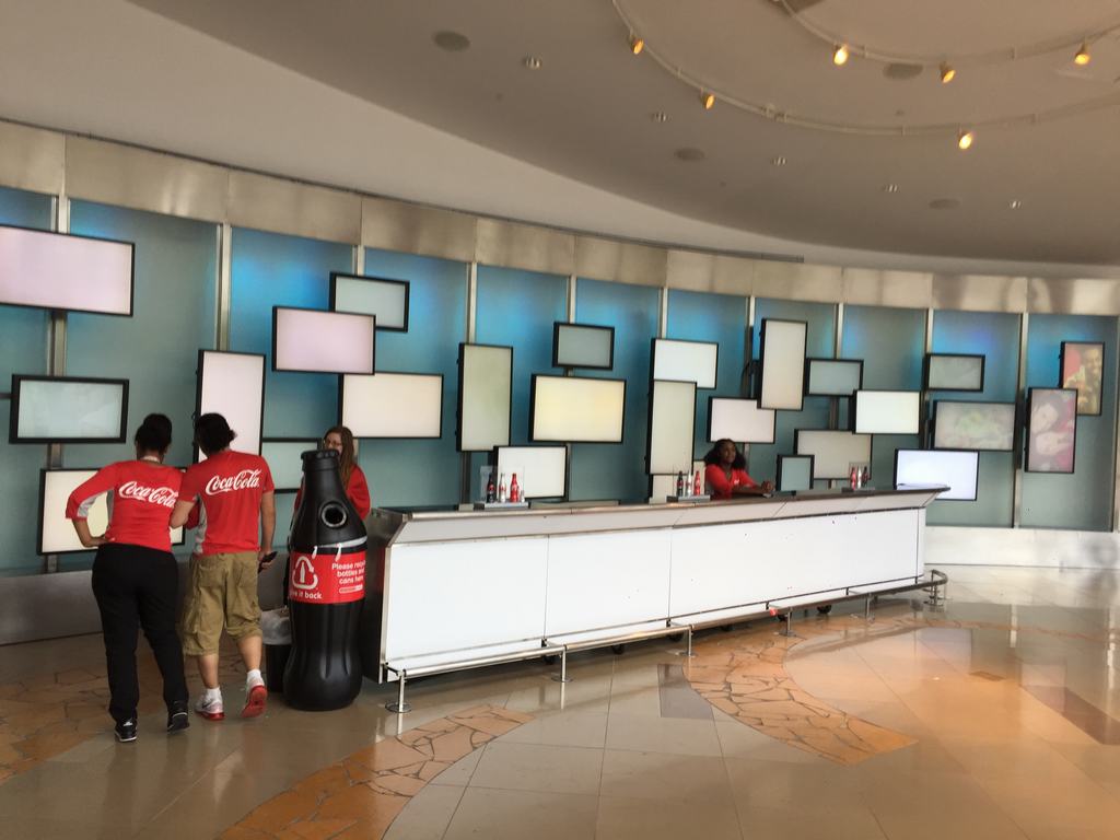 Obsluha v červených tričkách Coca-Cola u dlouhého pultu, stěna obrazovek