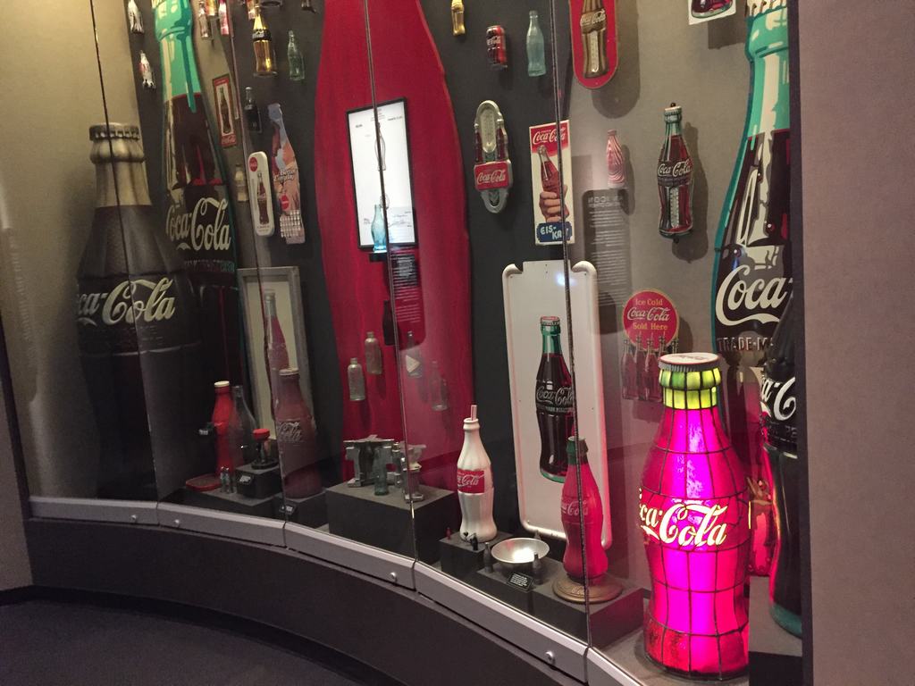Vitrína s historickými lahvemi a reklamami Coca-Cola, nápisy 5¢ a Ice Cold Coca‑Cola Sold Here.