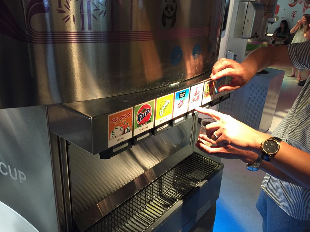 Návštěvník plní kelímek u automatu; volí příchuť s popisky JAPAN, THAILAND.