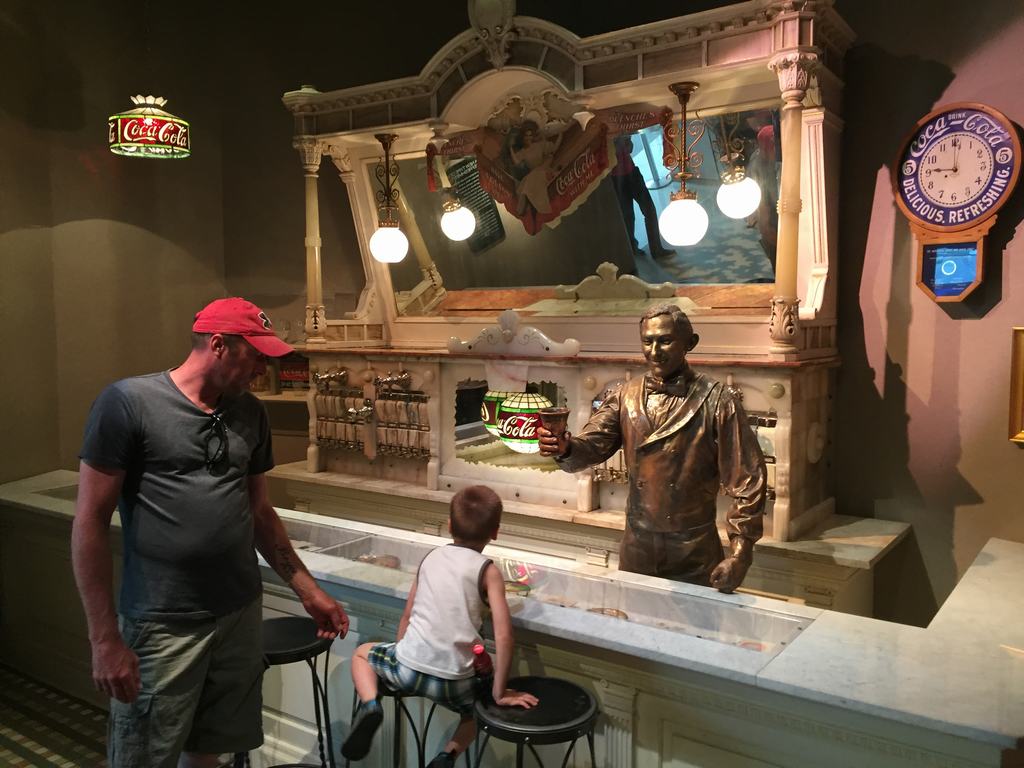 Muž a dítě u starého pultu, bronzová socha podává nápoj, Coca-Cola Delicious and Refreshing 5¢.