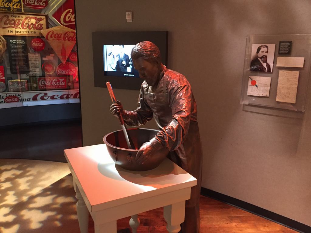 Socha muže míchajícího směs v míse; v pozadí nápisy Coca-Cola, DRINK Coca-Cola IN BOTTLES